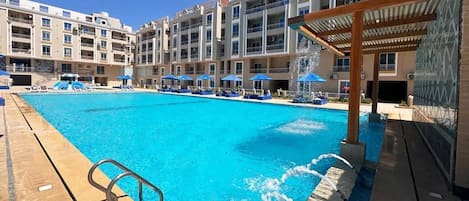 A swimming pool with a wooden deck, lounge chairs, and a covered area.
