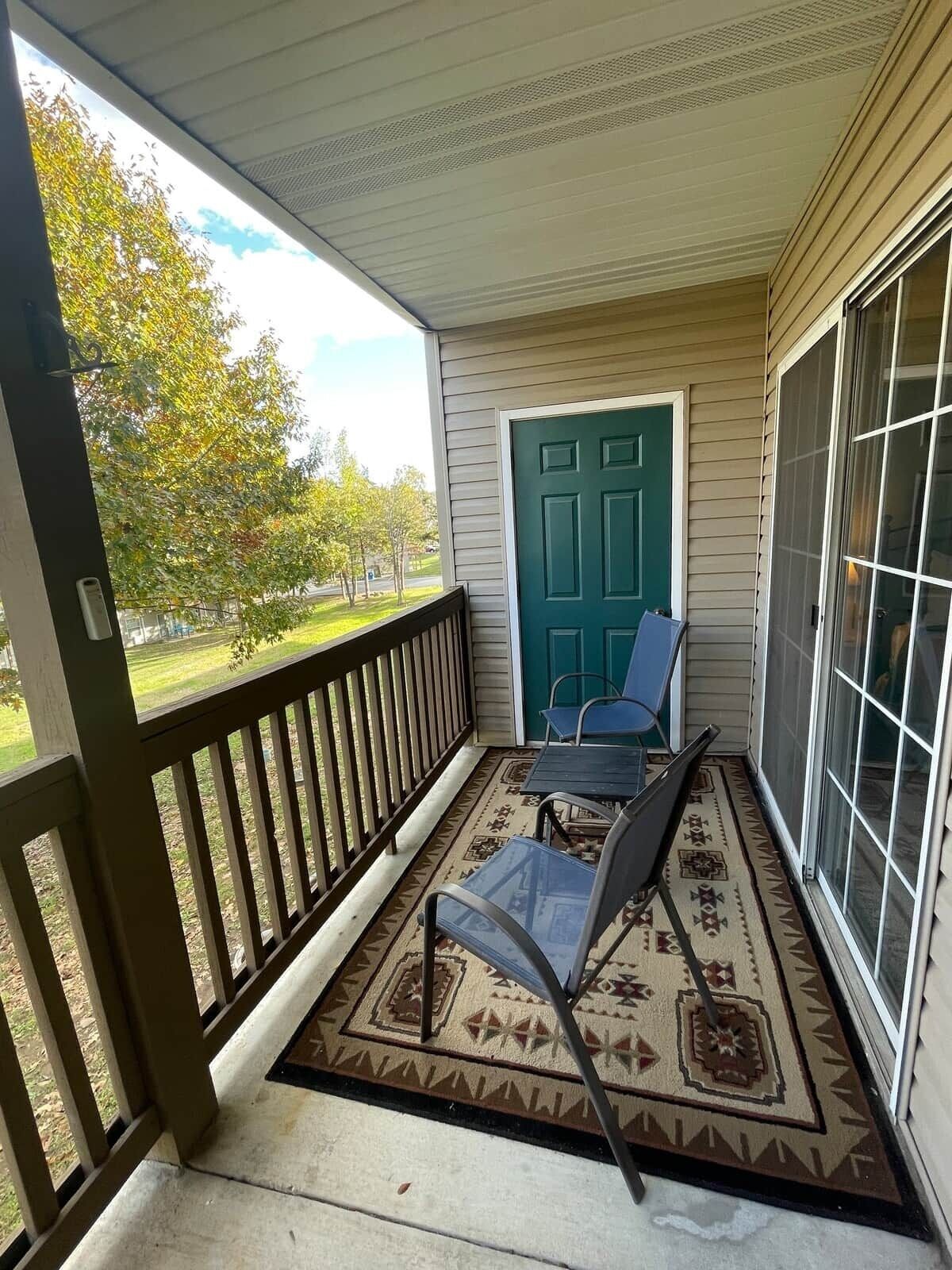 A covered porch with a view of a green door, a small table, and two chairs.