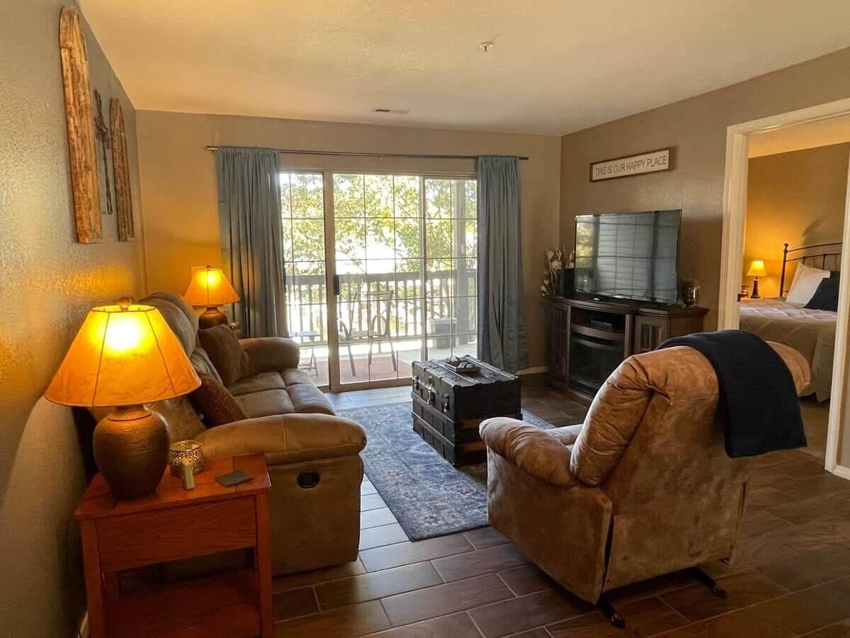 A living room with a sofa, armchairs, a coffee table, and a television. There is a sliding glass door leading to an outdoor area.