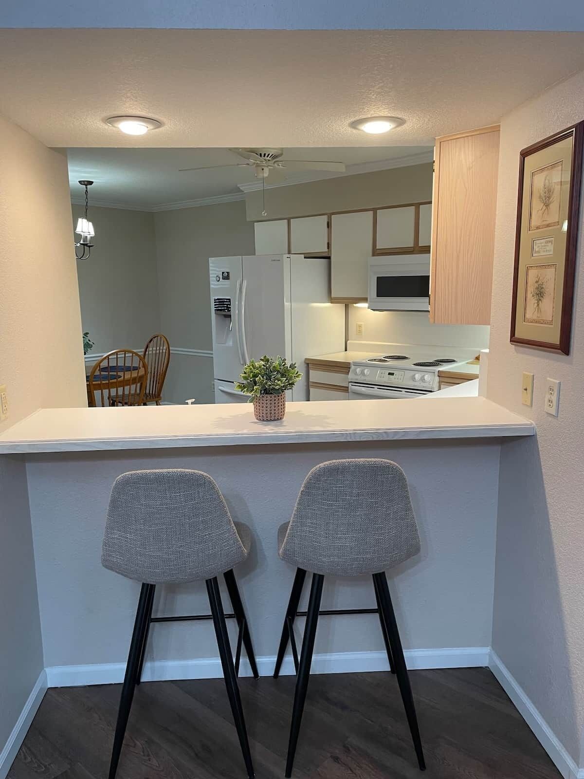 A modern kitchen with white cabinets, a refrigerator, a microwave, and a stove. There are two bar stools and a small potted plant on the counter.