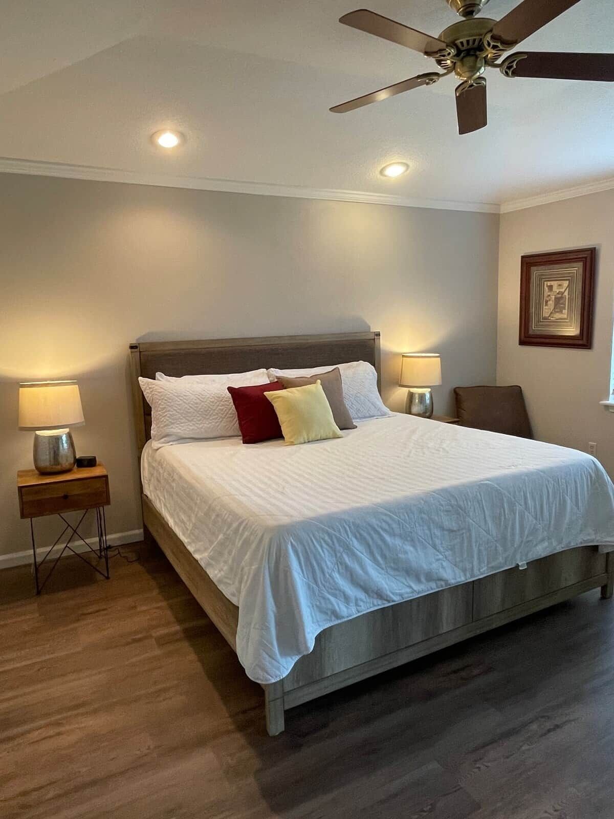 Une chambre à coucher avec un lit, deux tables de chevet avec des lampes, un ventilateur de plafond et un tableau encadré au mur.