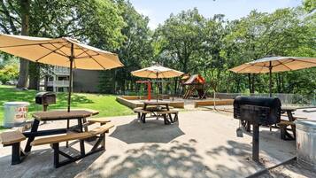 An outdoor patio area with picnic tables and umbrellas, surrounded by trees and a playground.