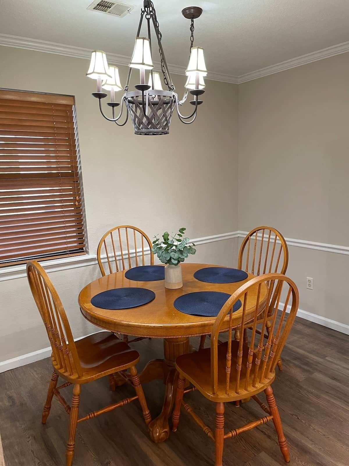 A dining area with a round wooden table, four wooden chairs, a chandelier, and a window with blinds.