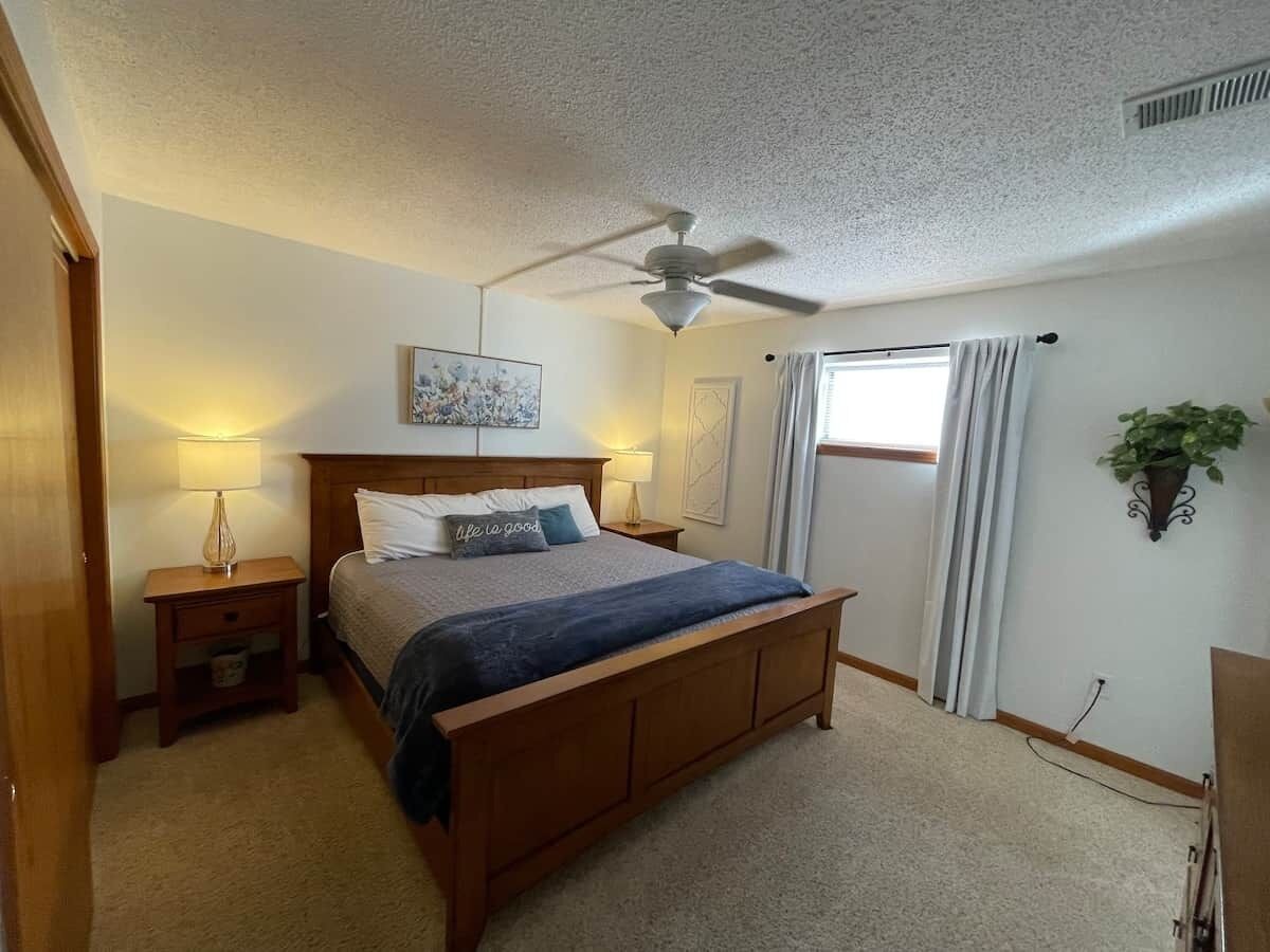 A bedroom with a bed, bedside tables, a ceiling fan, a window with curtains, and a wall-mounted plant.