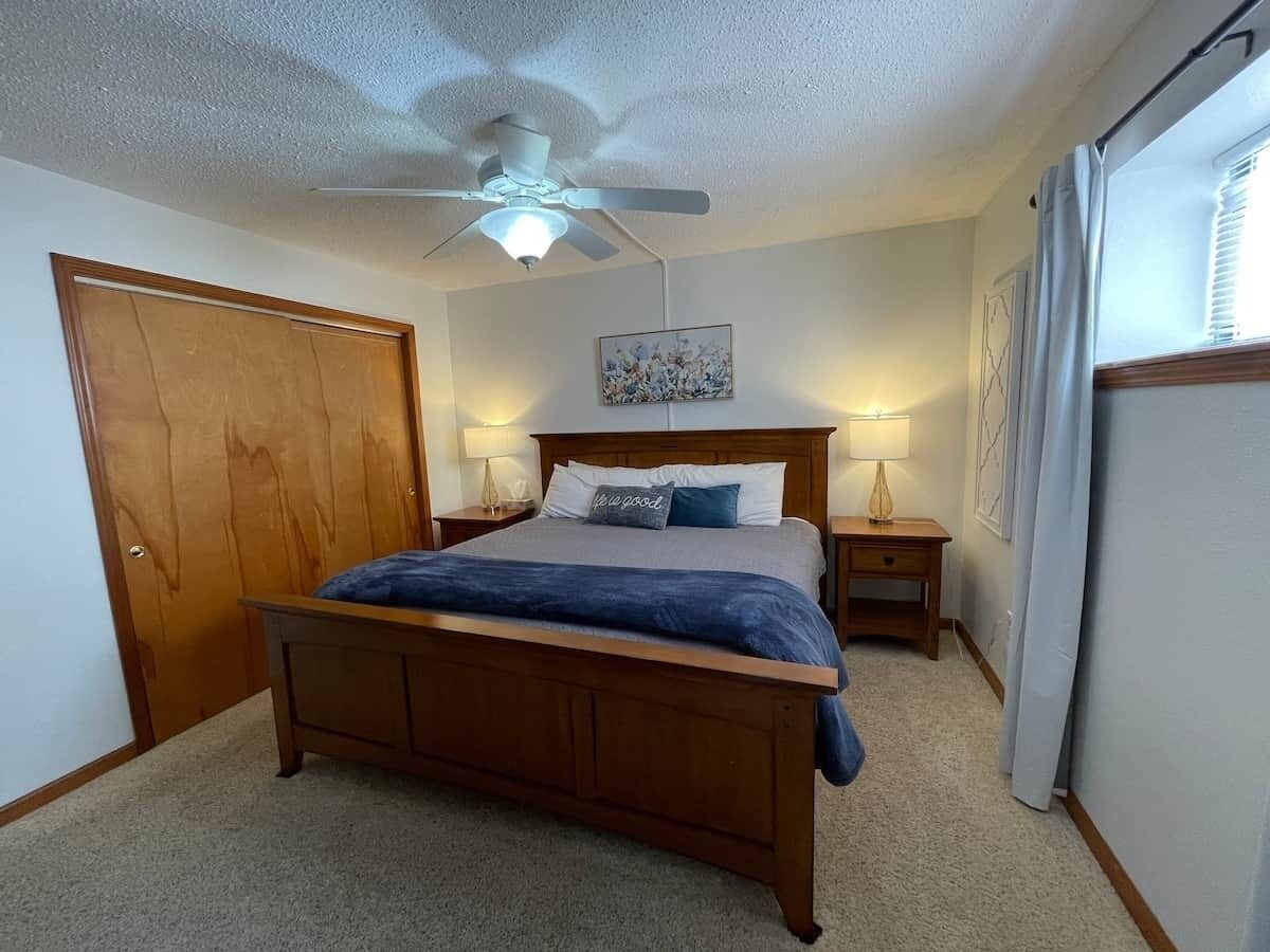 A bedroom with a wooden bed, a ceiling fan, two bedside tables with lamps, a door, and a window with curtains.