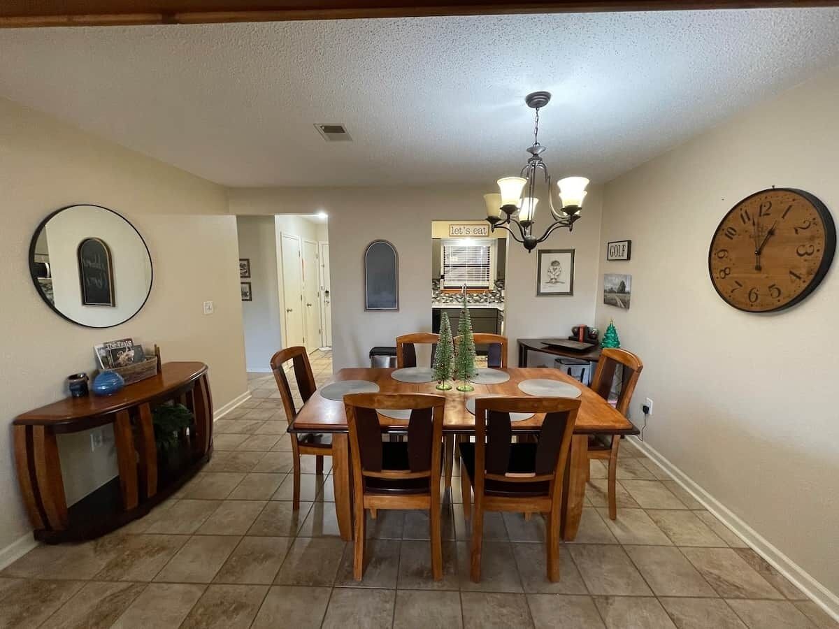 A dining area with a wooden table and chairs, a chandelier, a clock, and a sign that says 'let's eat.'