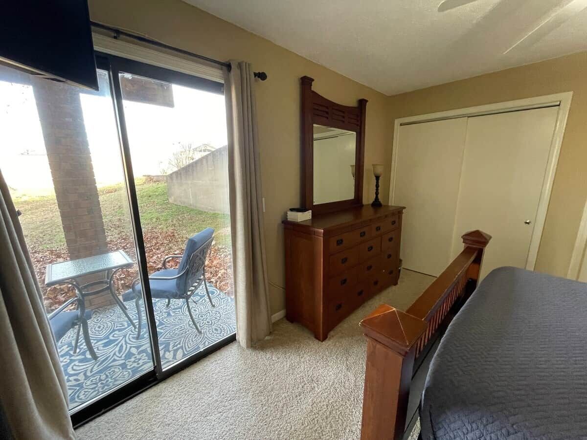 A bedroom with a bed, a dresser, a mirror, and a sliding glass door leading to an outdoor patio with a table and chairs.