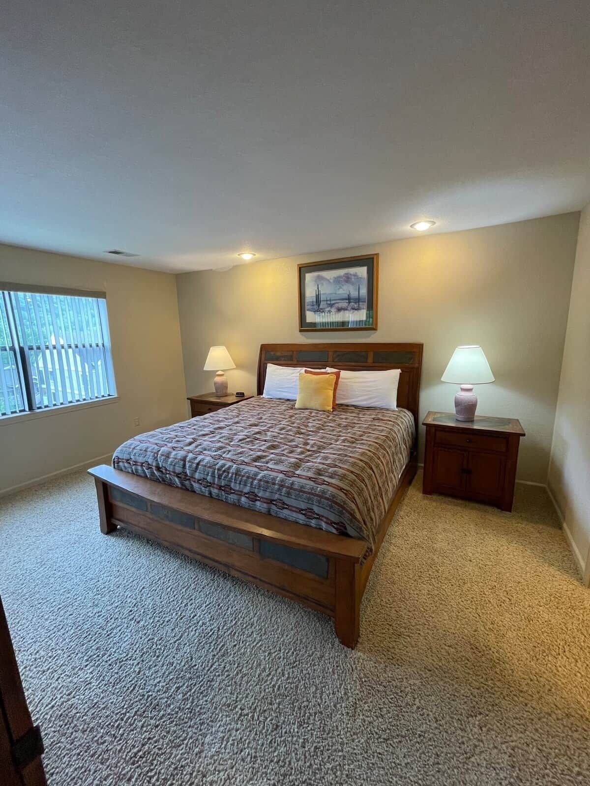 Une chambre à coucher avec un grand lit, deux tables de chevet avec des lampes, une fenêtre avec des stores et un tableau au mur.