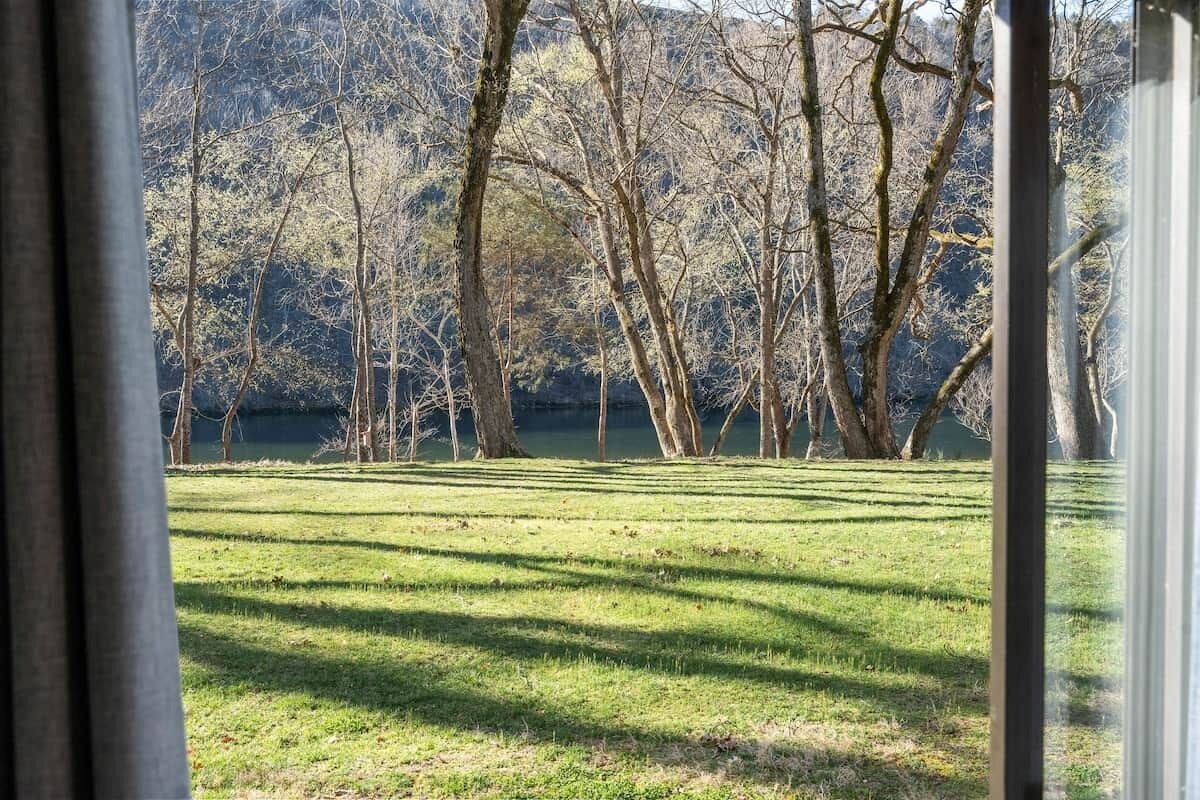 Une vue à travers une fenêtre sur une zone herbeuse, avec des arbres et une étendue d’eau en arrière-plan.