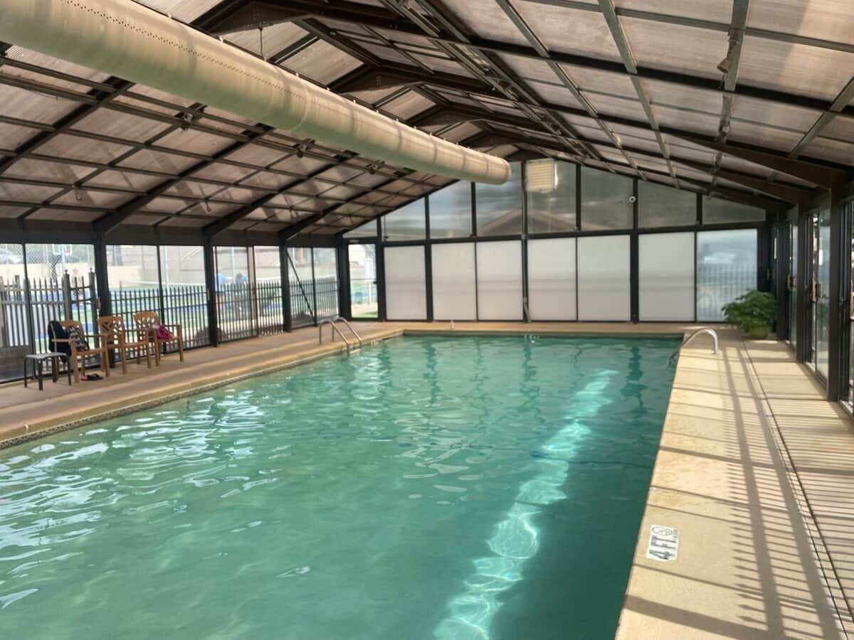 An indoor swimming pool with a clear water surface, surrounded by a covered area with a metal roof and glass walls.
