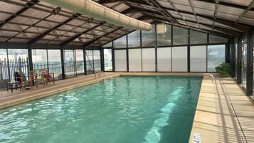 An indoor swimming pool with a clear water surface, surrounded by a covered area with a metal roof and glass walls.