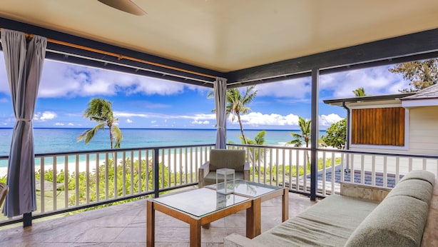 Varanda com mesa de vidro e sofá, com vista para uma praia com palmeiras e o oceano.