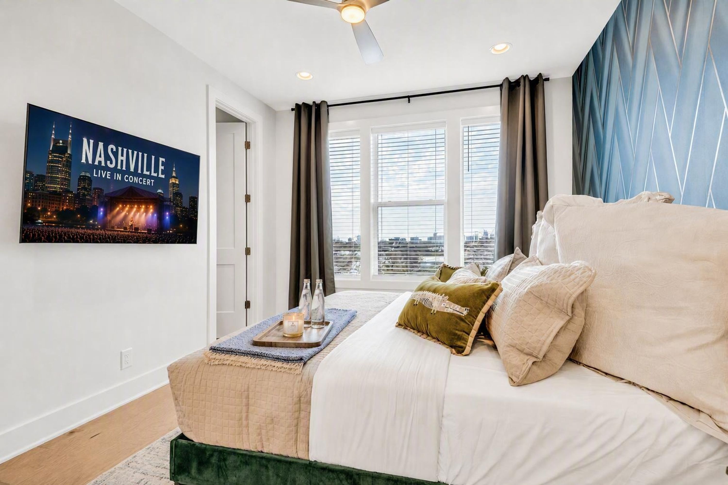 Una habitación de hotel con cama, una ventana con cortinas, un cartel con un paisaje urbano y una bandeja con velas.