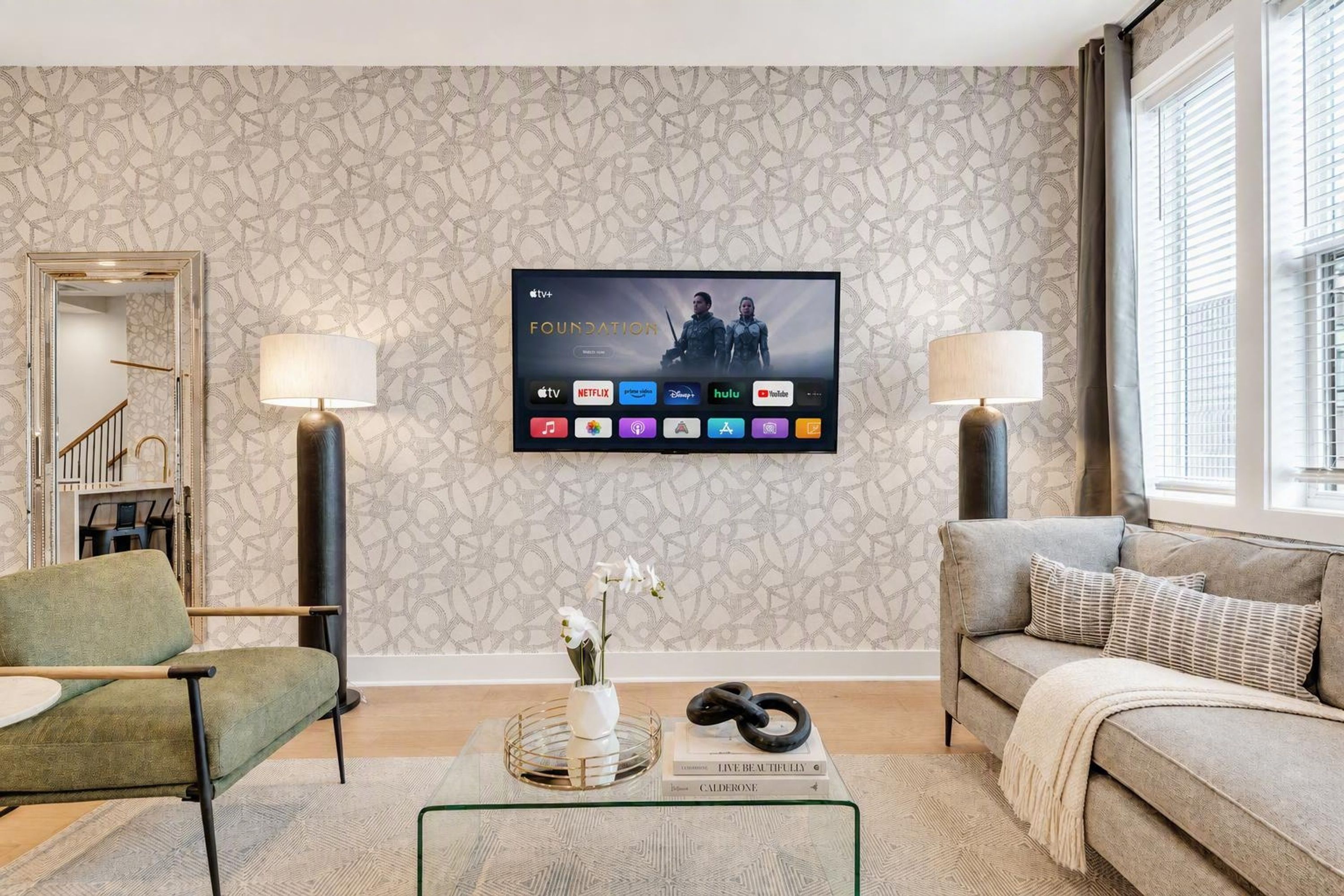 Una sala de estar con un televisor montado mostrando un programa, un sofá, una silla, una mesa de centro con objetos decorativos y un papel tapiz estampado.