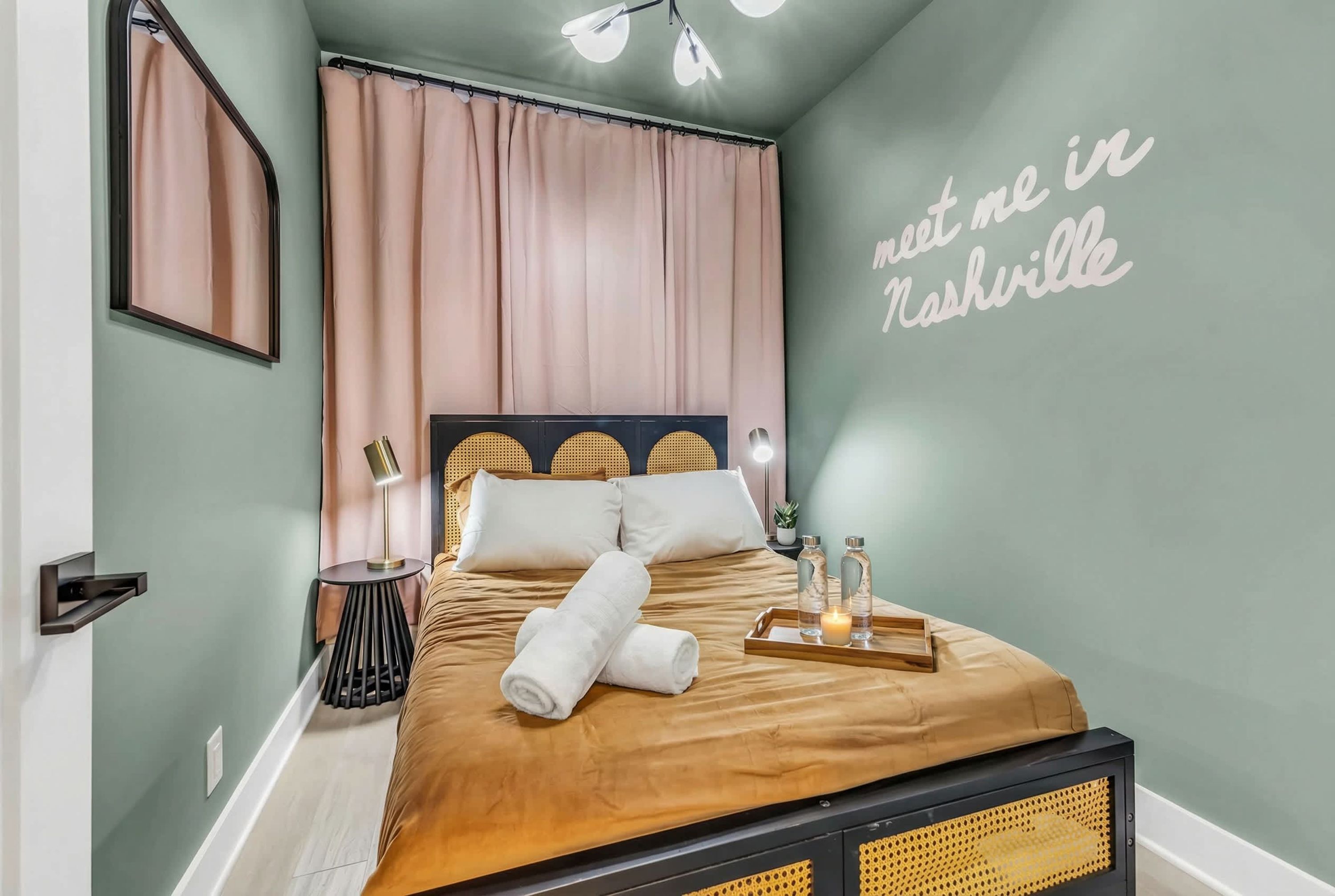 A neatly made bed with a brown cover, a bedside table with a lamp, a tray with bottles, and a wall with 'meet me in Nashville' text.