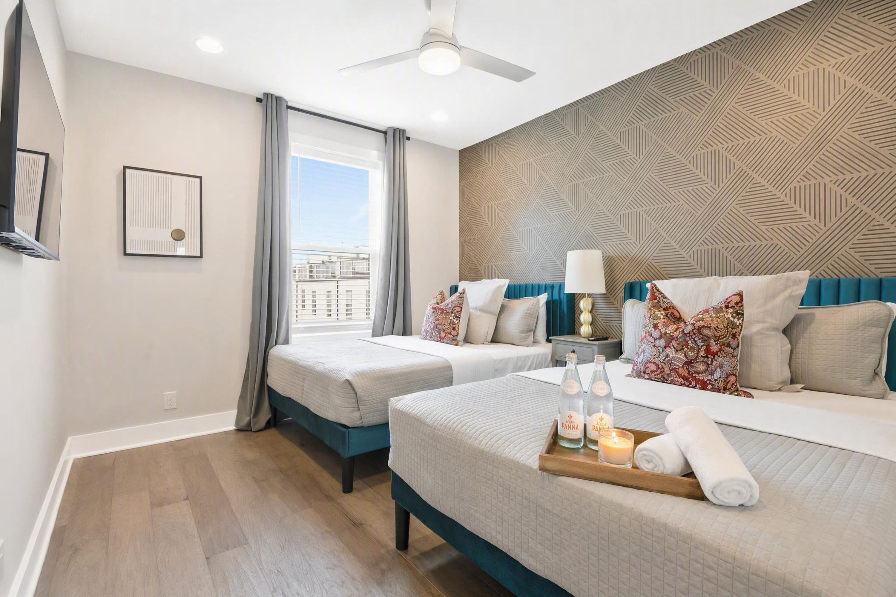 Habitación de hotel con dos camas, ventilador de techo, pared con diseño y ventana con cortinas.