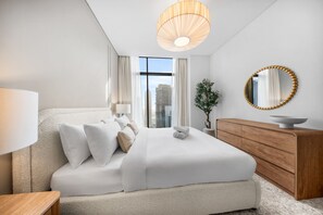 A modern bedroom with a large bed, a wooden dresser, a round mirror, and a view of the cityscape through the window. - Stayshort - 2 Bedroom in Business Bay Sleeps 6 With Water Canal Views (Dubai)