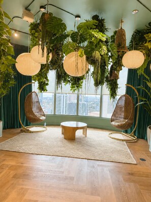 A room with a round wooden table, two wicker chairs, hanging plants, and a view of the city through large windows. - 501 First | StayChic Miami | Rooftop Pool & More (Miami)