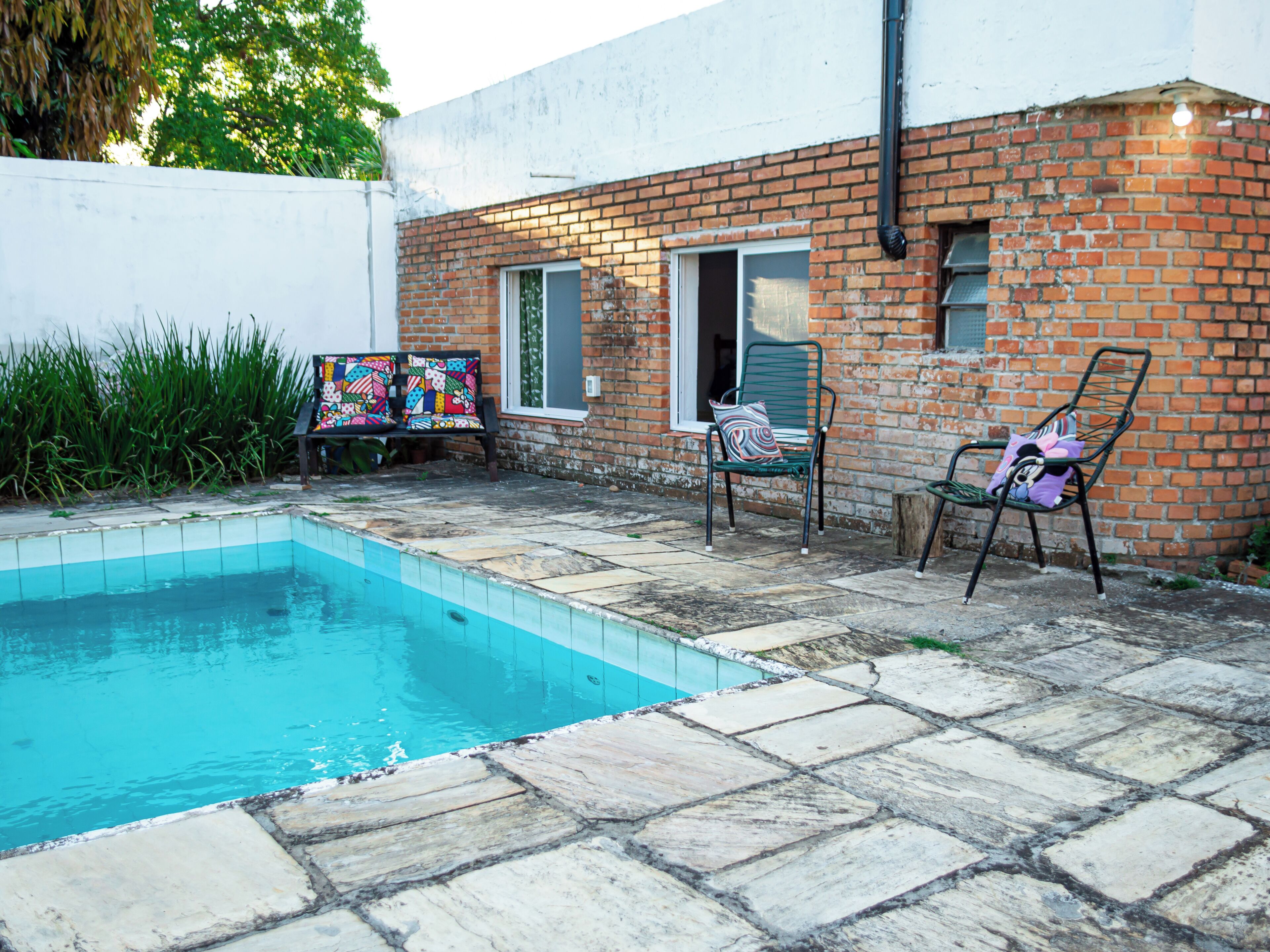 A swimming pool with stone tiles, two metal chairs with cushions, and a brick wall with a window.
