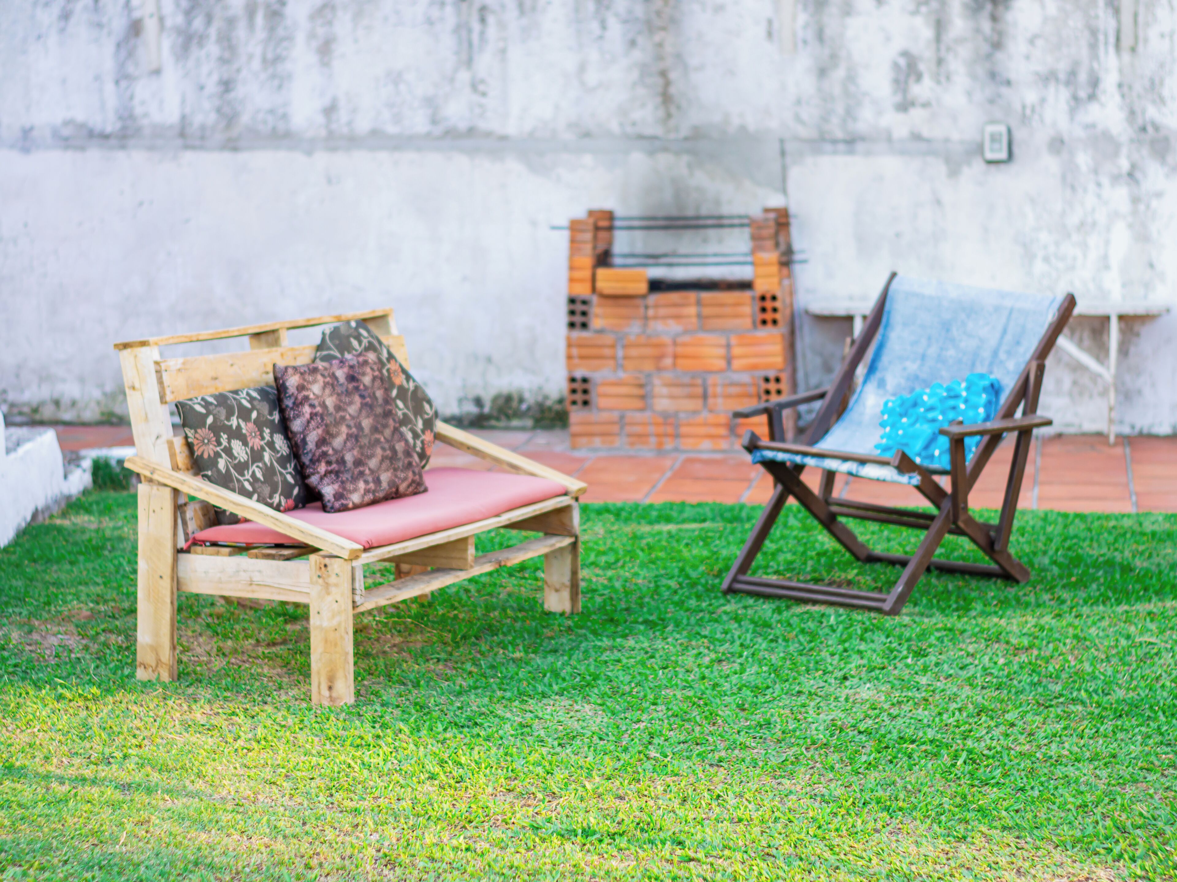 A wooden bench with cushions and a metal chair with a blue cushion in a grassy outdoor area.