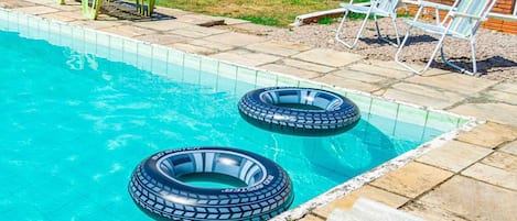 A swimming pool with two inflatable rings, a patio with chairs, and a brick building in the background.