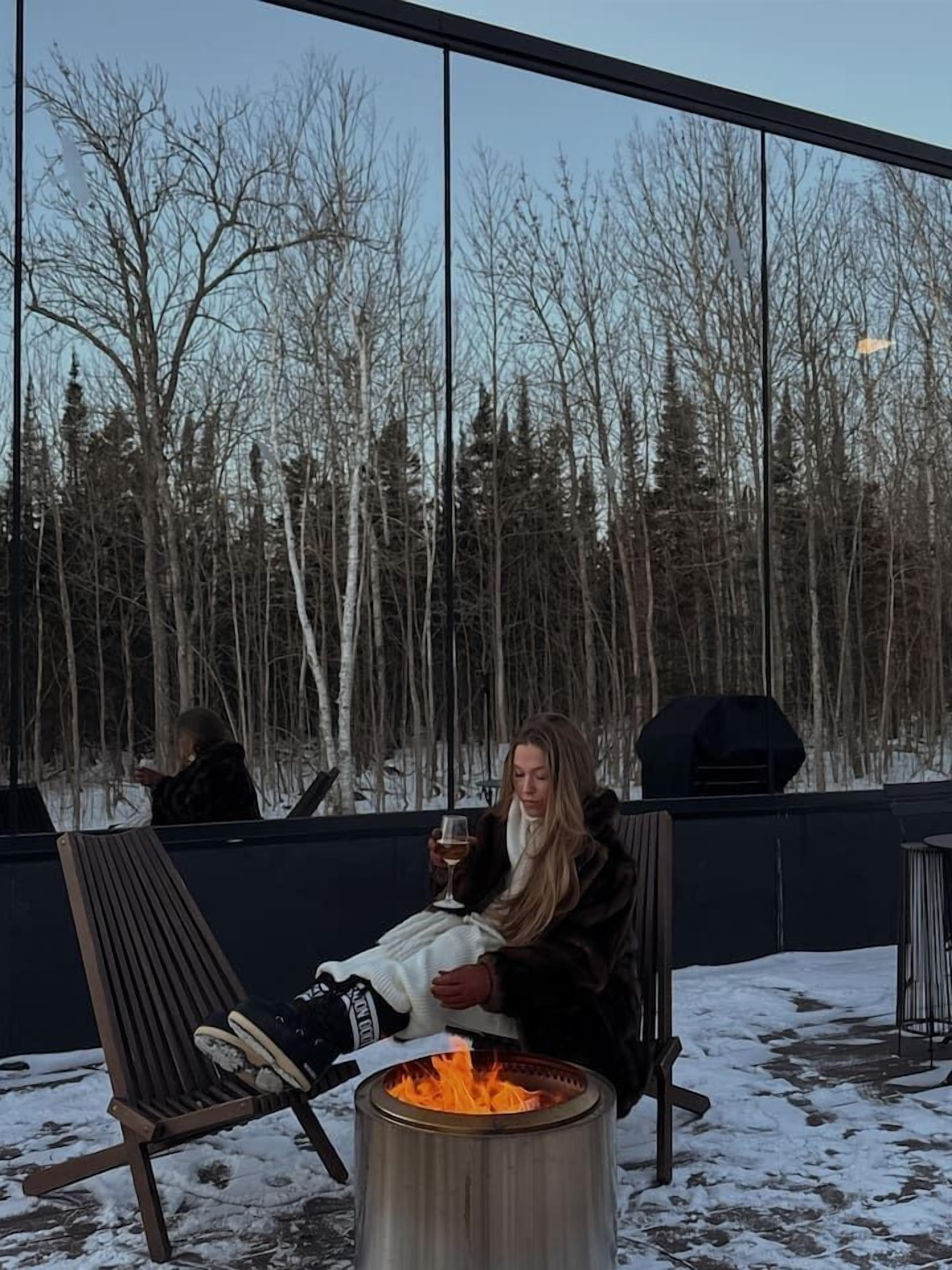 Henkilö istuu nuotiopaikan ääressä, ja hänellä on näkymä metsäiselle alueelle.
