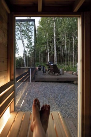 A wooden deck with outdoor furniture, a fire pit, and a view of a forest. - Aura | Mirror Cabin w/ Private Sauna (Lutsen)