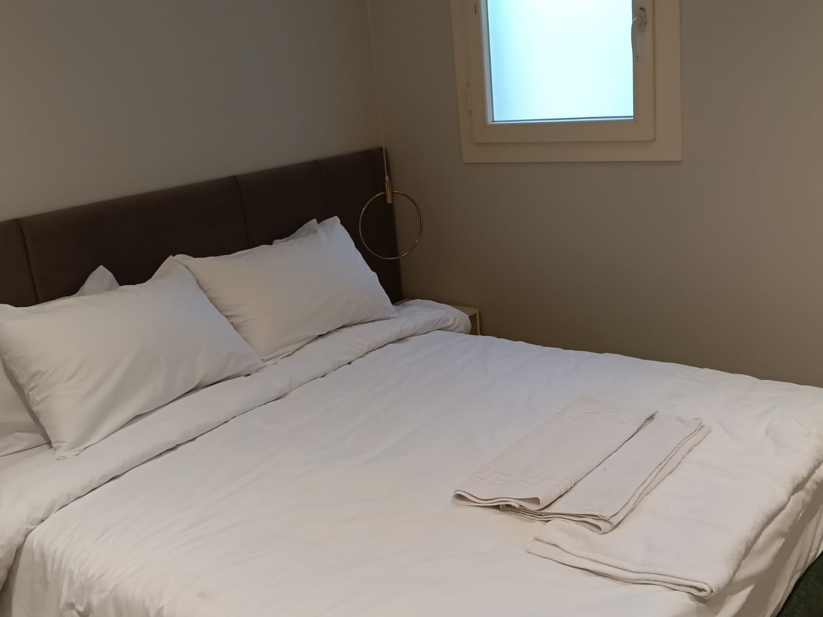 A neatly made bed with white linens, a headboard, and a window with daylight coming through.