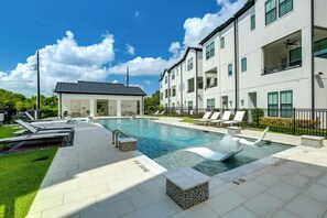 A modern pool area with lounge chairs, a small building, and a multi-story residential building with balconies. - Work + Unwind Poolside! Nurse-ready Houston Rental (Houston)