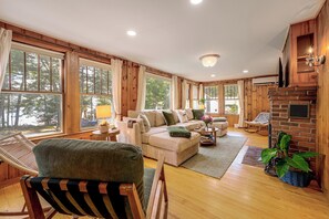 A cozy living room with a sofa, armchairs, a fireplace, and large windows. - Sebago Lake Cottage Boat Mooring 2 Beaches (Windham)