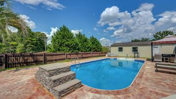 A swimming pool with a stone border, a wooden fence, and a covered barbecue area.