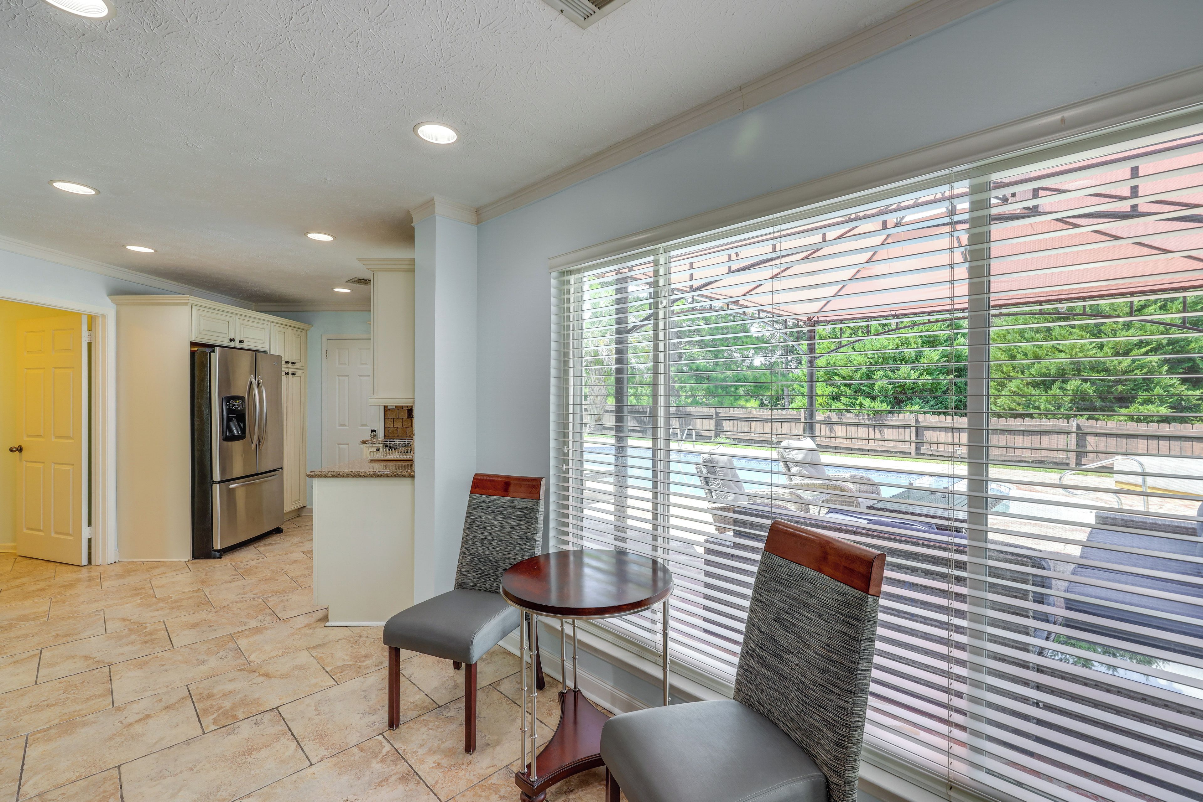 A kitchen with a stainless steel refrigerator, a dining area with two chairs and a table, and a large window with blinds.