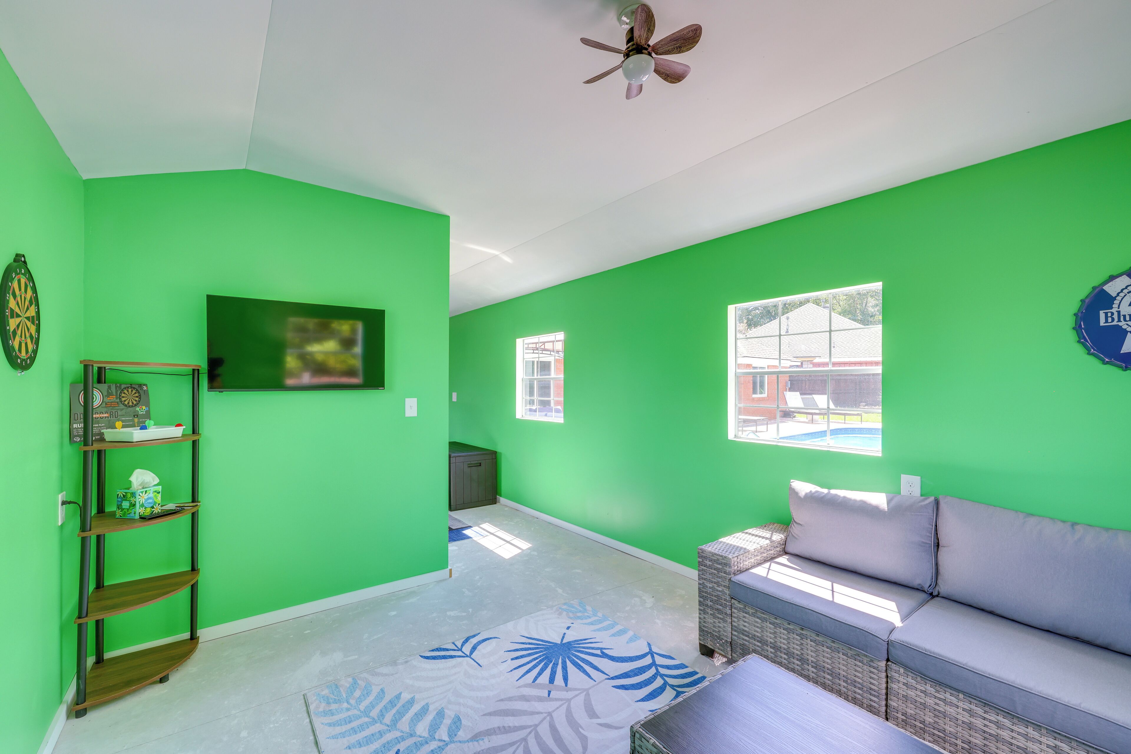 Une pièce aux murs verts, avec un ventilateur de plafond, un jeu de fléchettes, une fenêtre et un canapé.