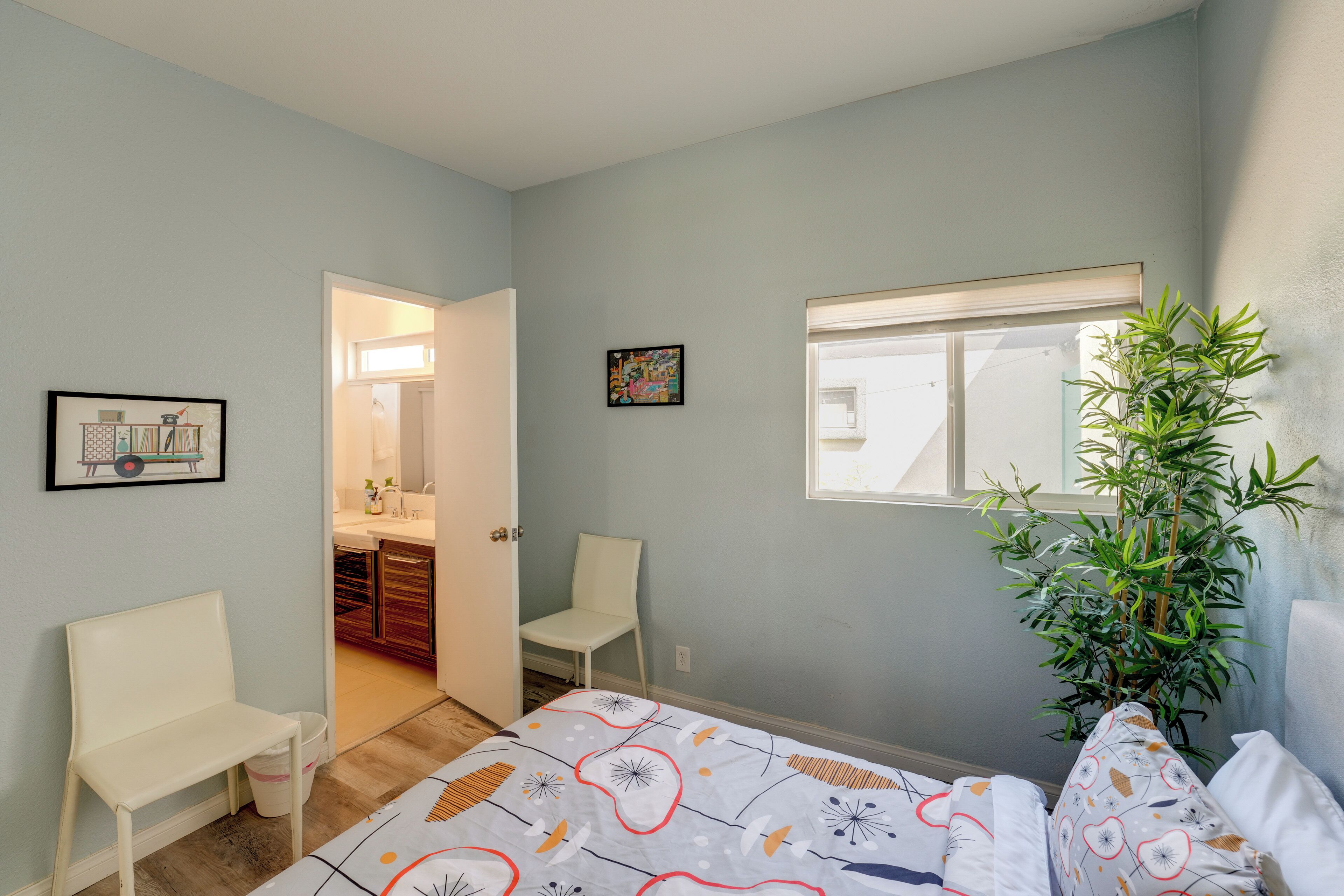 A bedroom with a bed, a chair, a plant, a window, and a door leading to a bathroom.