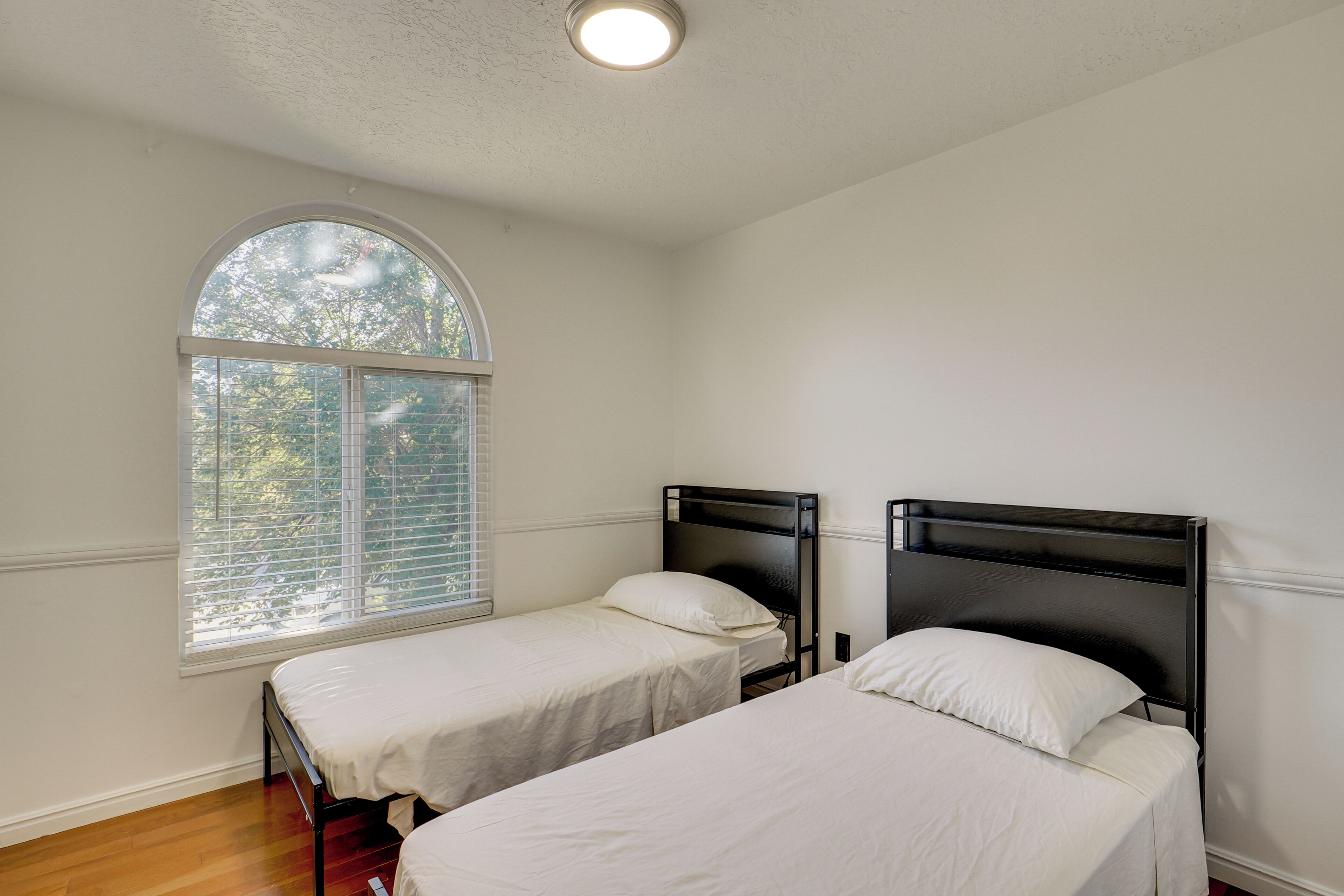 A room with two beds, a large arched window, and a ceiling light.