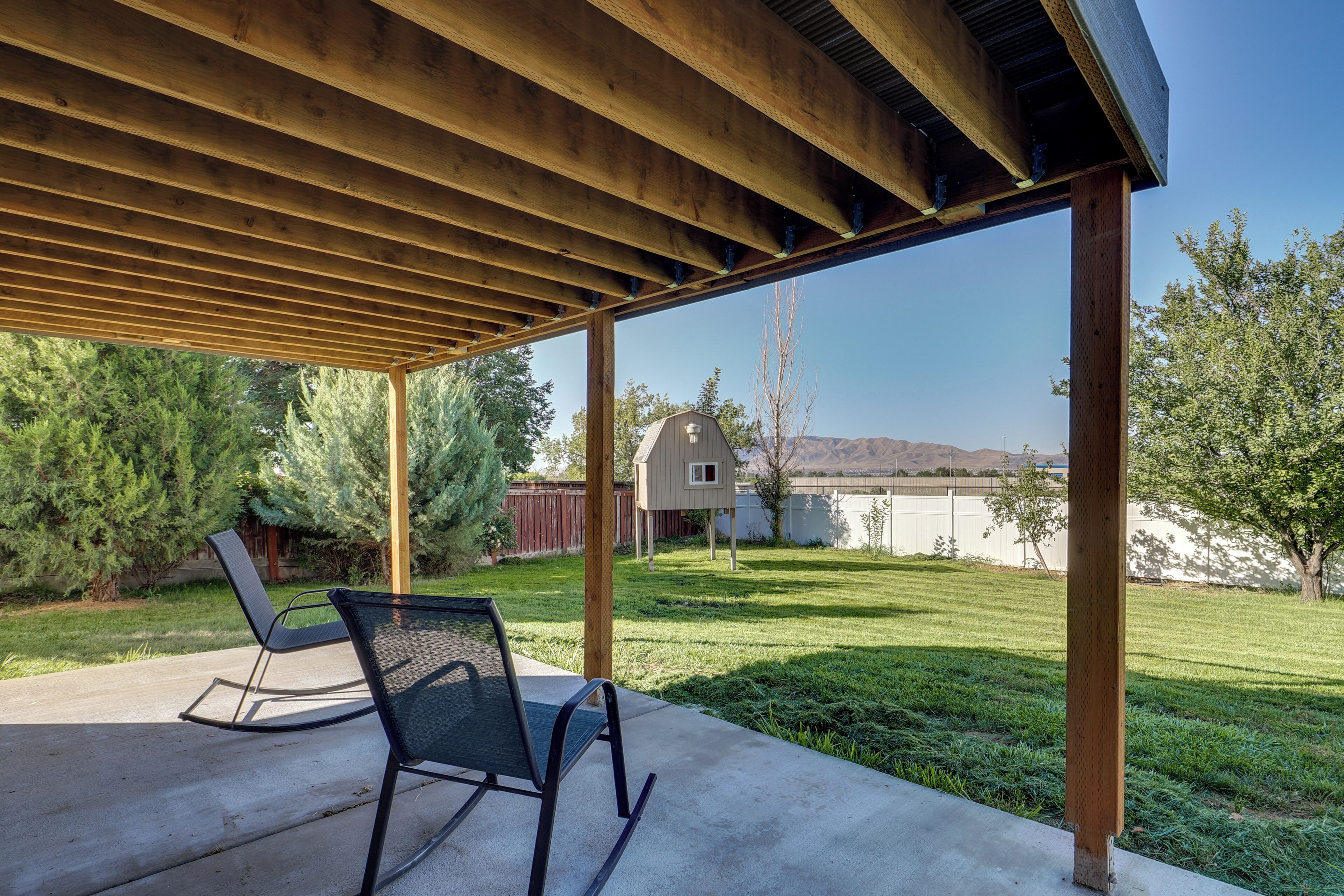 Un patio couvert comprenant deux chaises, une pergola en bois et une vue sur une cour arrière avec une maison et des montagnes au loin.