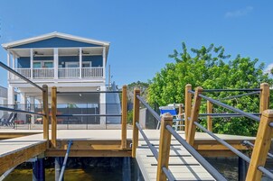 A wooden dock leading to a house with a balcony, a clear sky, and a tree in the background.