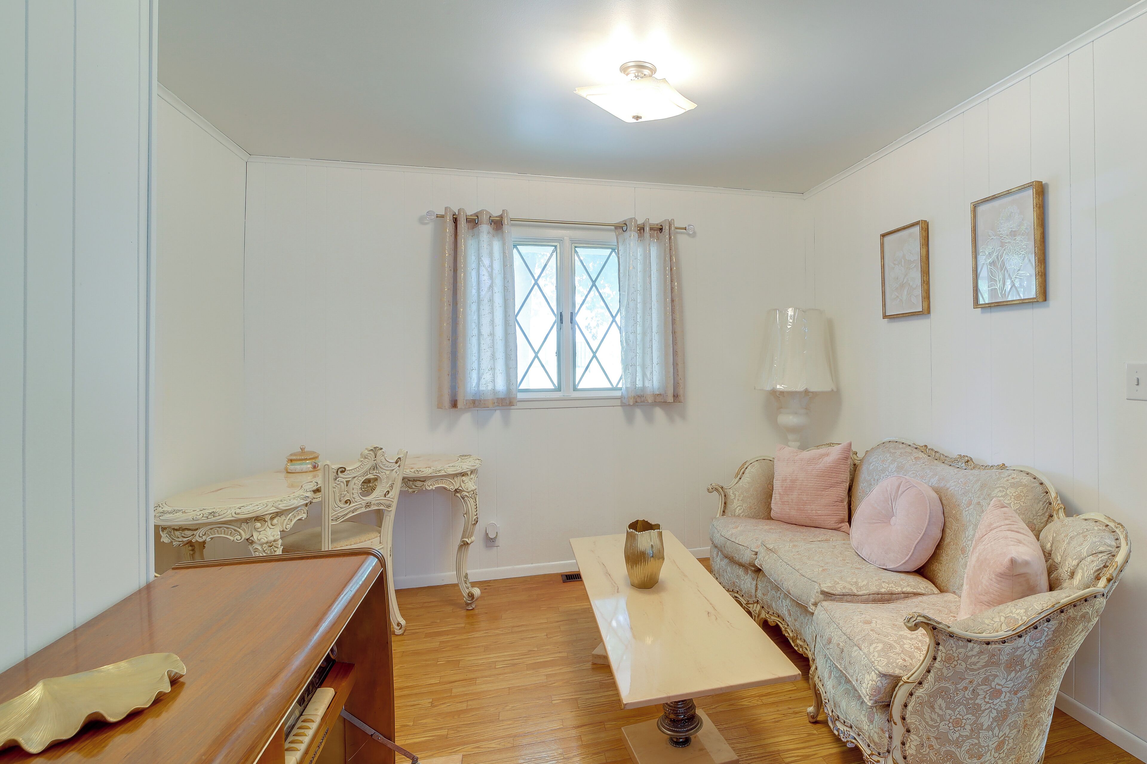 Una habitación con piso de madera, un escritorio blanco con silla, un sofá con cojines, una ventana con cortinas e iluminación en el techo.