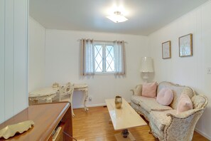 Una habitación con piso de madera, un escritorio blanco con silla, un sofá con cojines, una ventana con cortinas e iluminación en el techo.