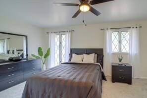 Ein Schlafzimmer mit einem Bett, einer Kommode, einem Nachttisch, einem Deckenventilator und zwei mit Vorhängen versehenen Fenstern.