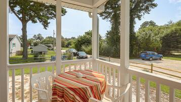 A covered porch with a table and chairs, overlooking a street with parked cars and trees.