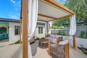 Ein überdachter Außenbereich mit Pergola, Korbmöbeln und Blick auf ein Haus im Hintergrund.