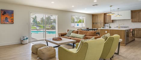 A spacious living area with a pool view, wooden flooring, and a modern kitchen.