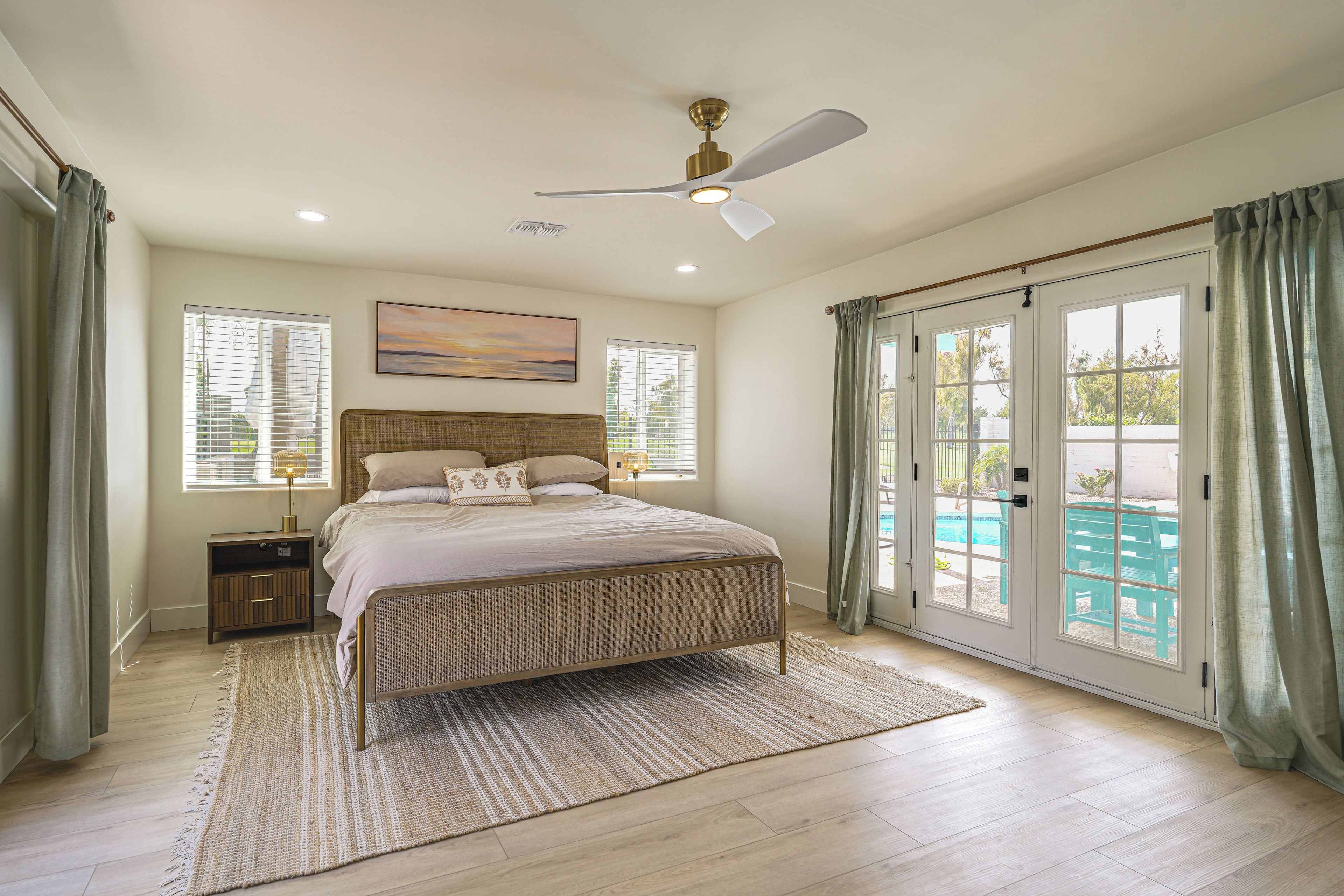 Quarto com cama, mesa de cabeceira, ventilador de teto e vista para uma piscina através das portas de vidro.