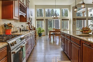 A spacious kitchen with wooden cabinets, a large island, and a view of the forest through the windows. - Exquisite Private Home Retreat Near Kenai River (Soldotna)