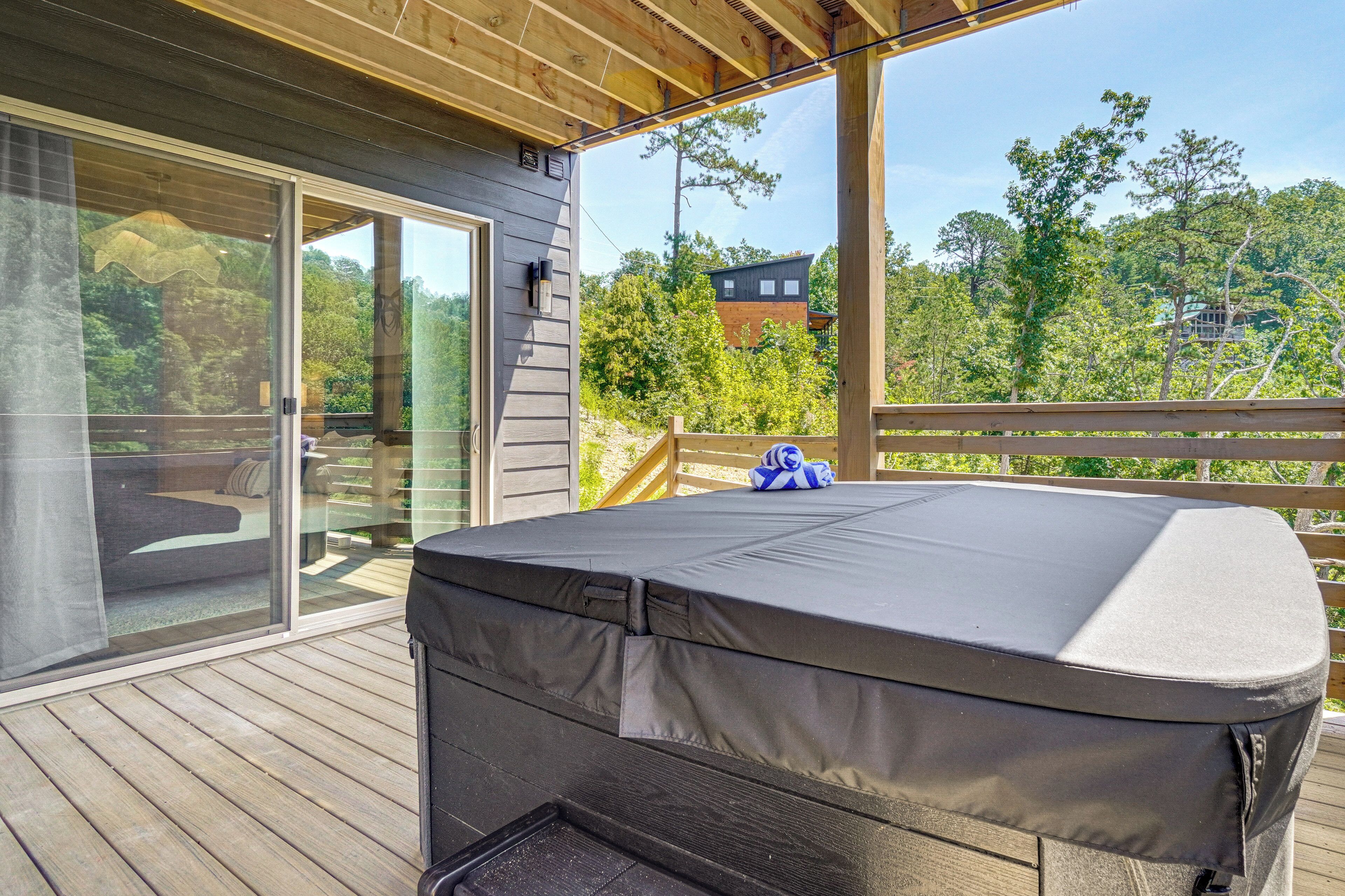 A covered outdoor deck with a hot tub, glass doors leading to a patio, and a view of surrounding trees and buildings.