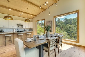 A modern kitchen with wooden cabinets, a large island with bar stools, and a dining area with a view of the forest. - Brand-new Smokies Cabin w/ Spectacular Mtn Views (Sevierville)