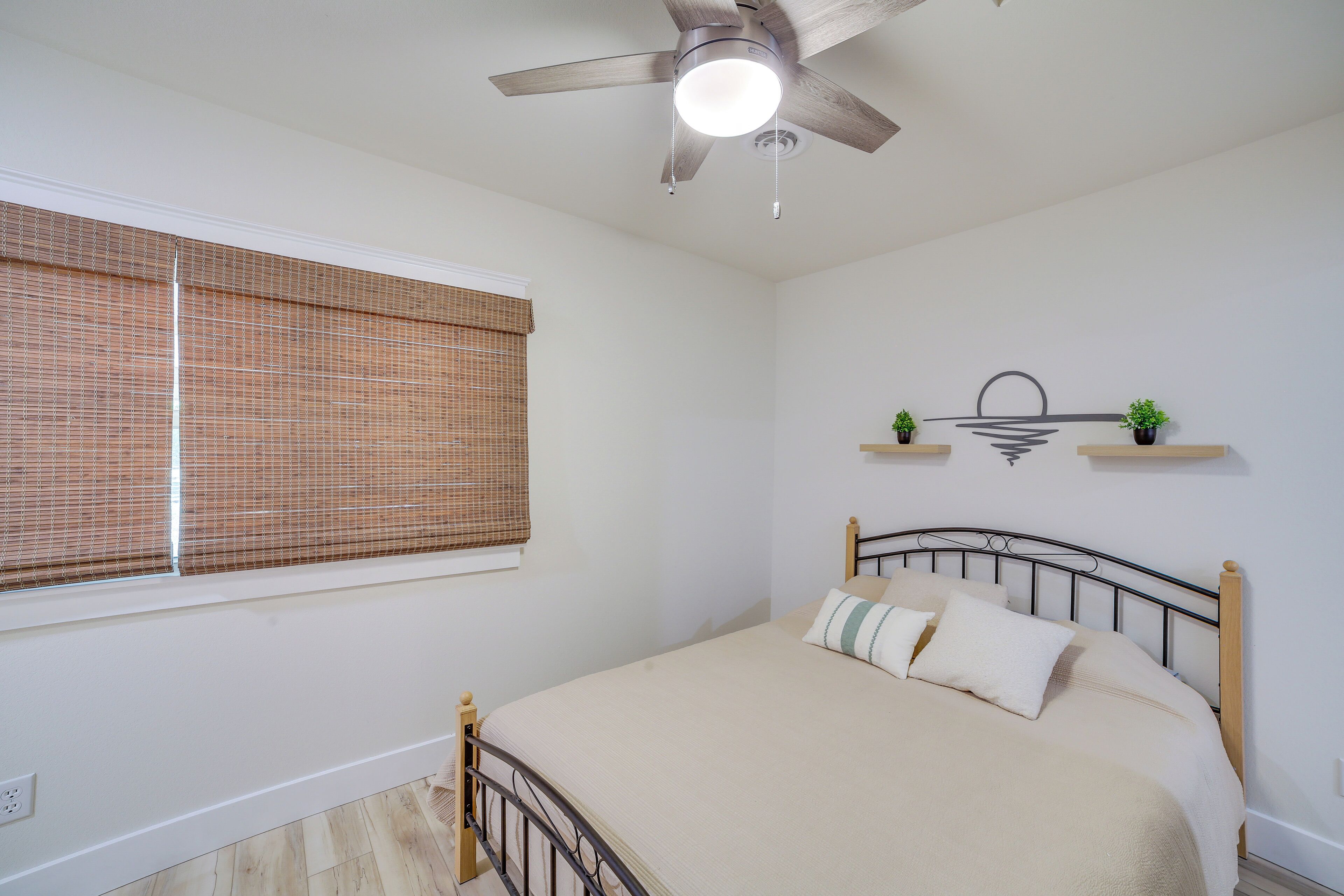 A bedroom with a bed, a ceiling fan, a window with blinds, and wall-mounted shelves with plants.