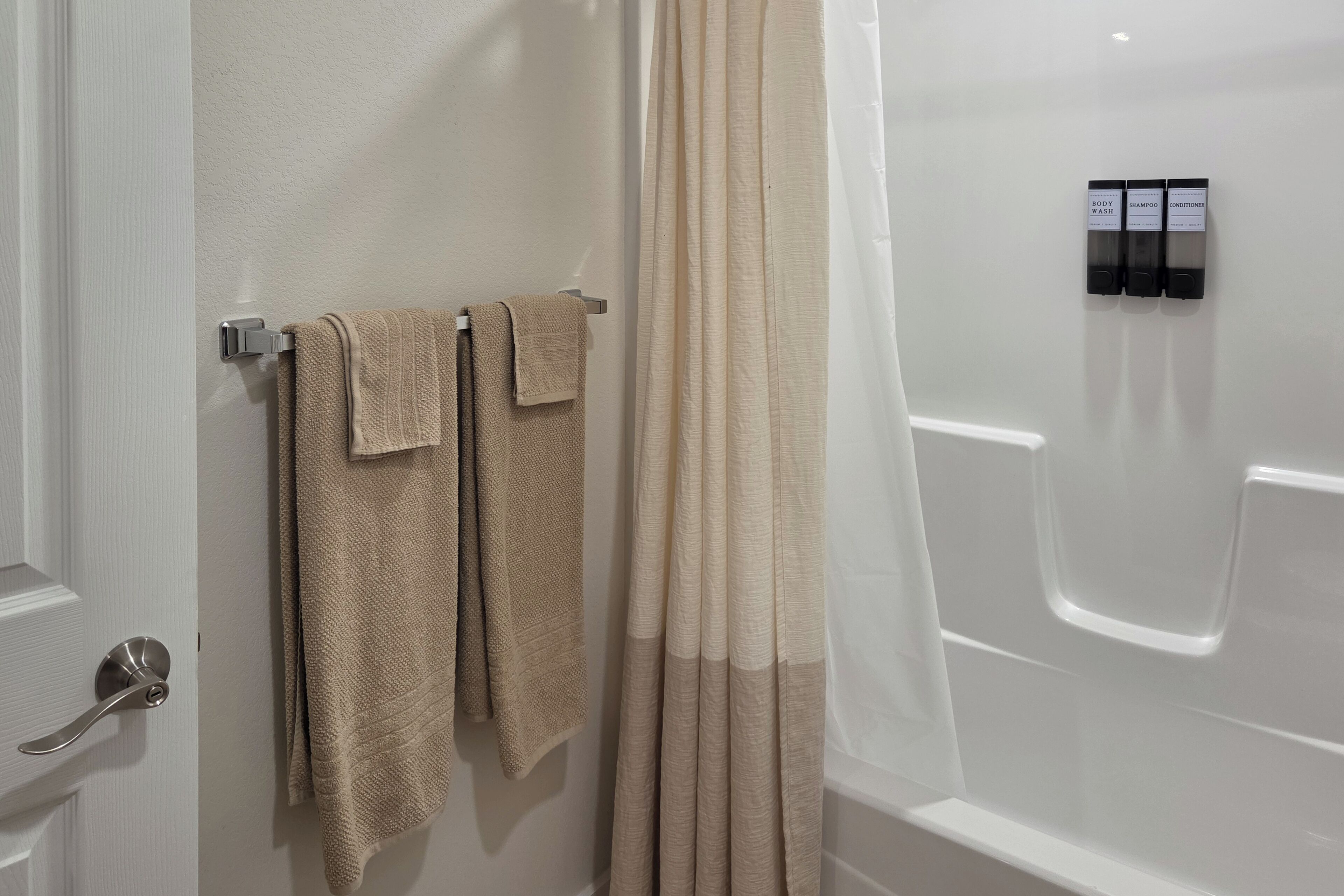 Une salle de bain avec une baignoire, un rideau de douche et un porte-serviettes avec deux serviettes. On y trouve également trois flacons étiquetés « gel douche », « shampoing » et « après-shampoing », qui sont accrochés au mur.