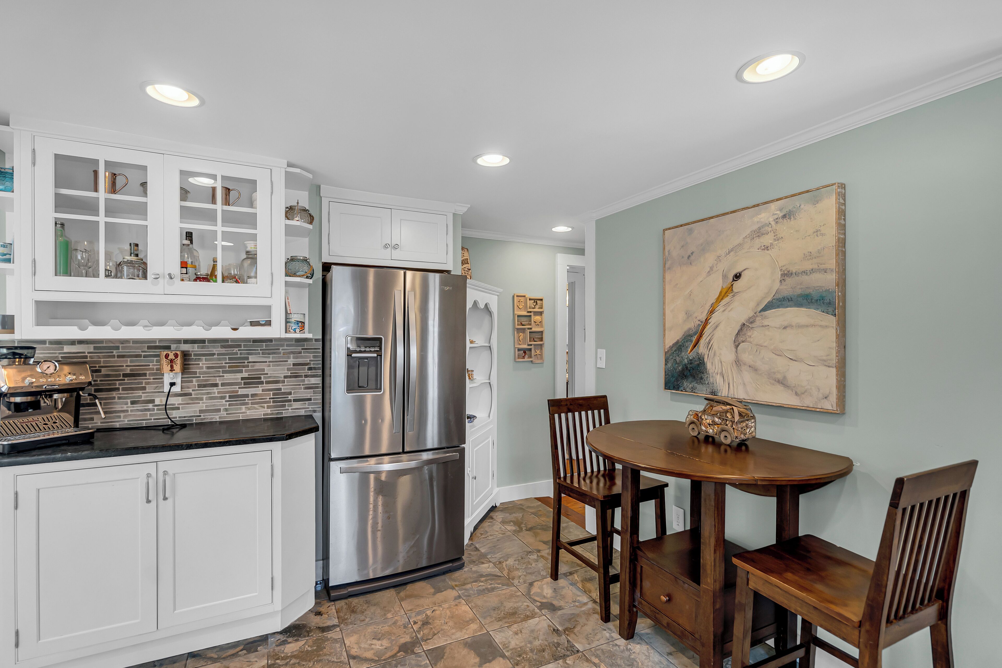 A kitchen with white cabinetry, a stainless steel refrigerator, a coffee machine, and a dining area with a wooden table and chairs.